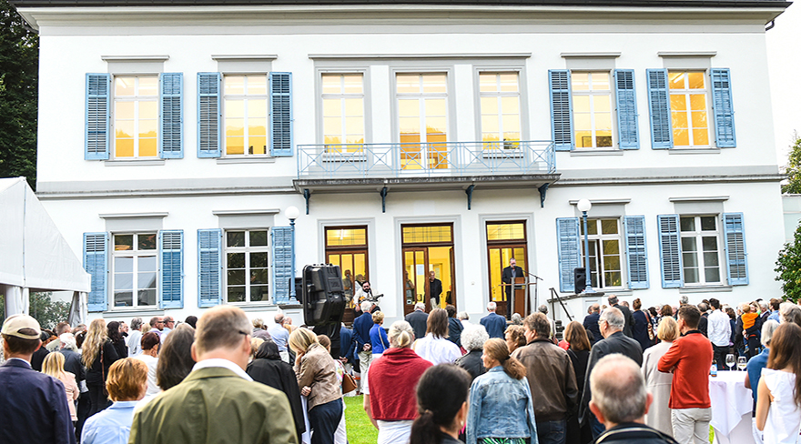 Auf diesem Bild sieht man eine Menschenmenge vor einem weißen Gebäude mit blauen Fensterläden. Die Leute stehen im Freien und blicken auf das Gebäude, wo scheinbar eine Veranstaltung stattfindet. Die Fenster des Gebäudes leuchten warmes Licht aus.