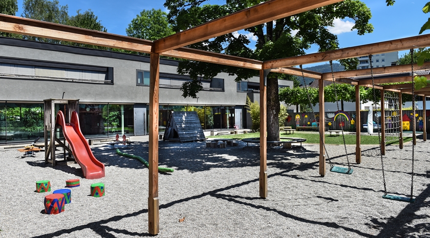 Hier wurde der Außenbereich des Kindergartens Braike in Bregenz fotografiert. Vor dem Gebäude sind Schaukeln, eine Rutsche und Klettergerüste zu sehen.