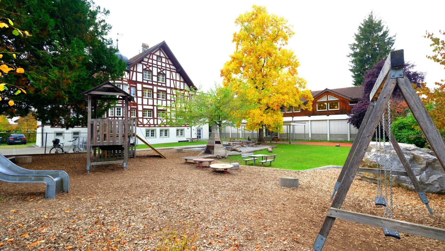 Hier wurde der Außenbereich des Kindergarten Weinschlössle fotografiert. In diesem finden sich eine Rutsche, Schaukeln, eine Kletter-Ecke, Sitzmöglichkeiten sowie zwei Bäume, wovon einer grün und der andere gelb blüht.