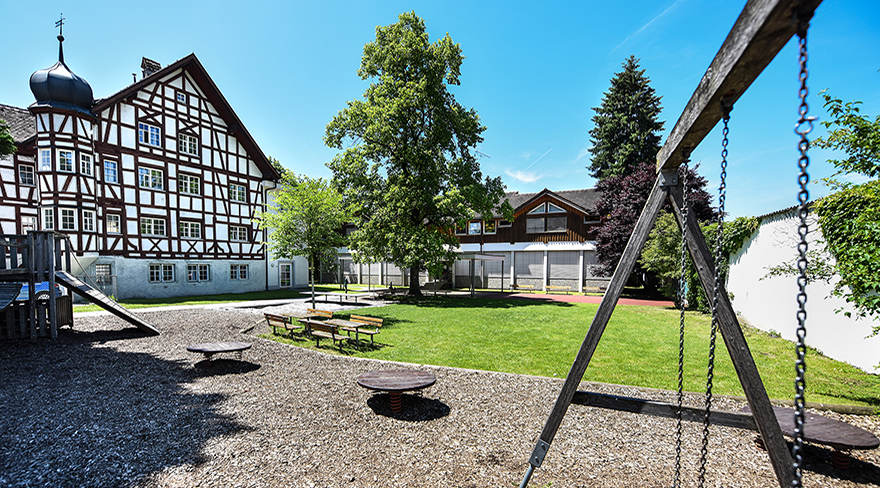 Diese Abbildung zeigt den Außenbereich des Kindergartens Weinschlössle in Bregenz. In diesem befinden sich eine Kletter-ecke, Schaukeln und diverse Sitzmöglichkeiten.