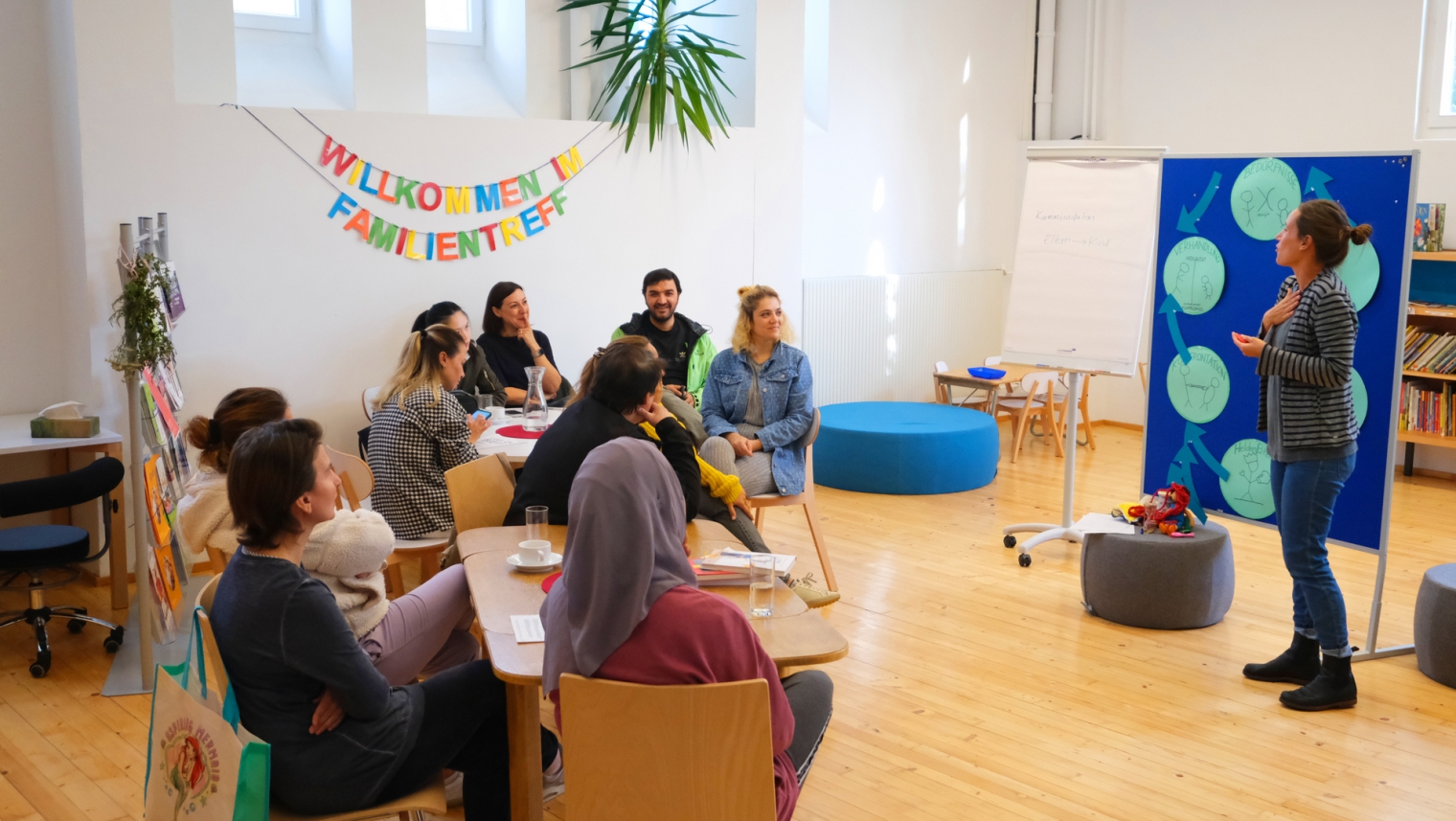 Auf der Abbildung sind mehrere Menschen auf Stühlen sitzend, sowie eine Vortagende, die im Familientreffpunkt ein Thema erörtert, zu sehen. Auf der Wand steht in bunten Buchstaben "Willkommen im Familientreff".