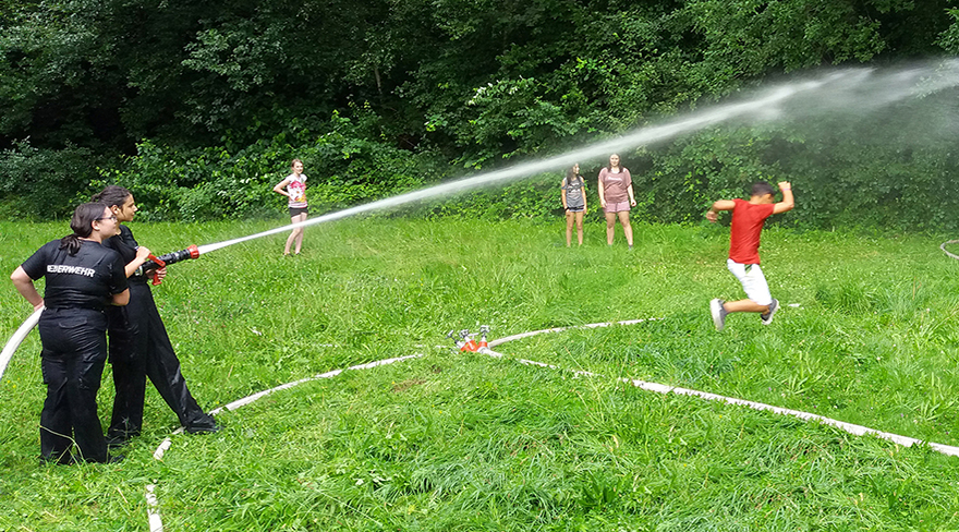 Diese Abbildung zeigt Kinder und Jugendliche, welche sich auf einer Wiese mit einem Wasserschlauch der Feuerwehr abkühlen.