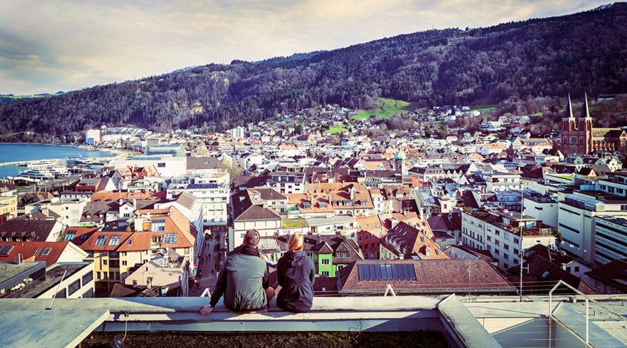 Diese Abbildung zeigt zwei Männer, welche auf einem Dach sitzen und einen Ausblick auf Bregenz haben.