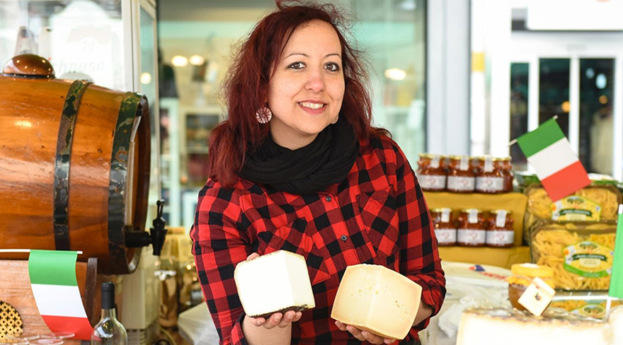 Diese Abbildung zeigt eine Dame, welche bei ihrem italienischen Stand zwei Stücke Käse in den Händen hält.