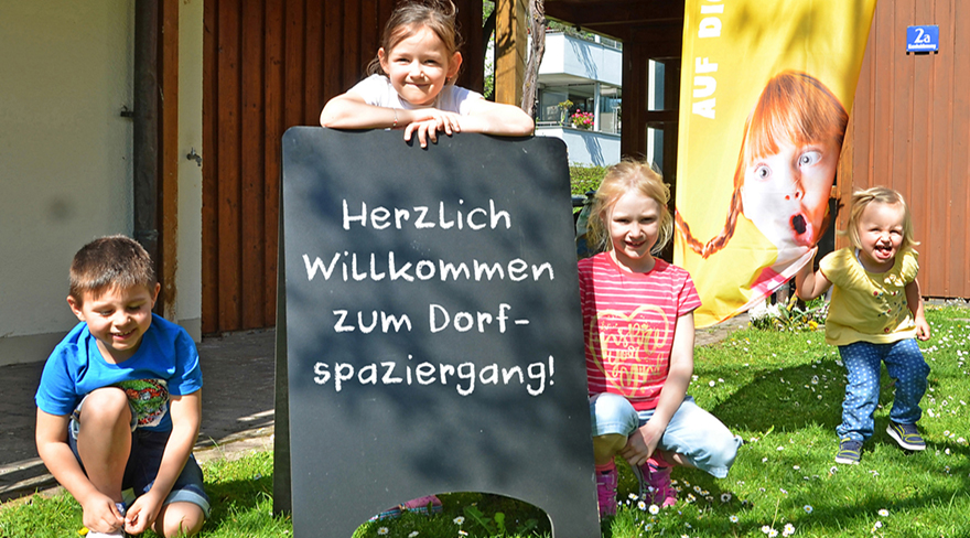 Diese Abbildung zeigt vier Kinder mit einer Tafel, auf welcher "Herzlich Willkommen zum Dorfspaziergang!" steht.