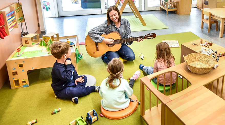 Diese Abbildung zeigt eine Pädagogin, welche den Kindern etwas auf der Gitarre vorspielt.